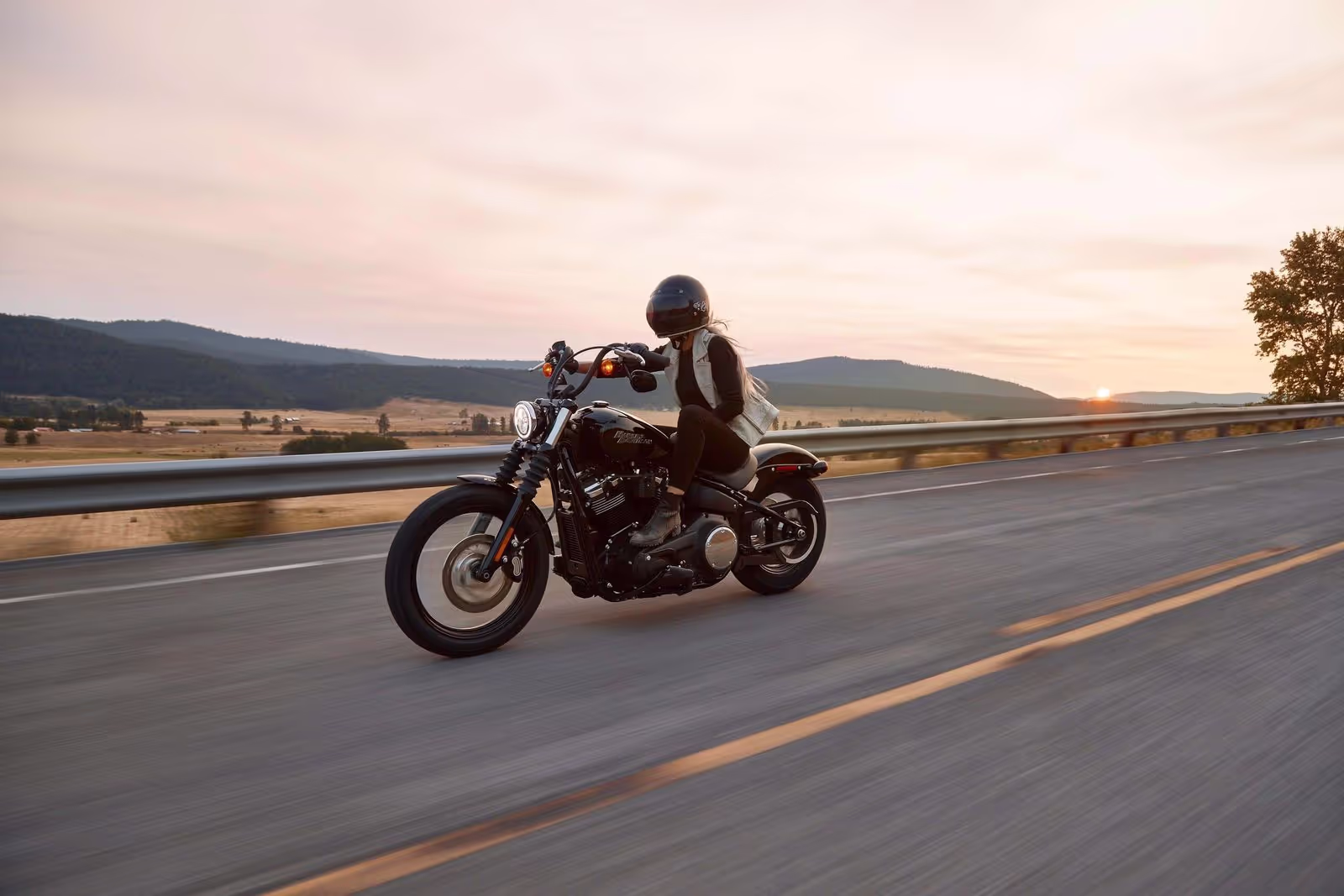 A woman rides a motorcycles down an open road with rolling hills in the background.